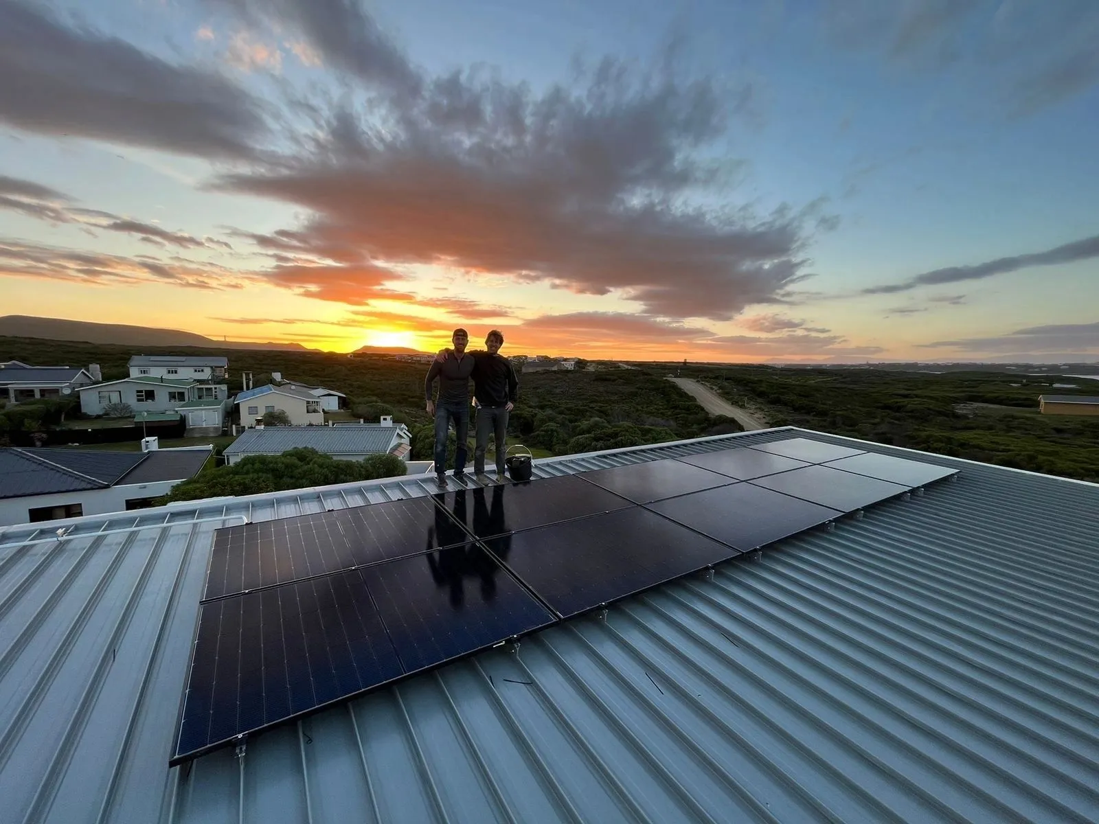Two installers standing on a metal roof beside a newly fitted solar array at sunset, coastal village behind them.