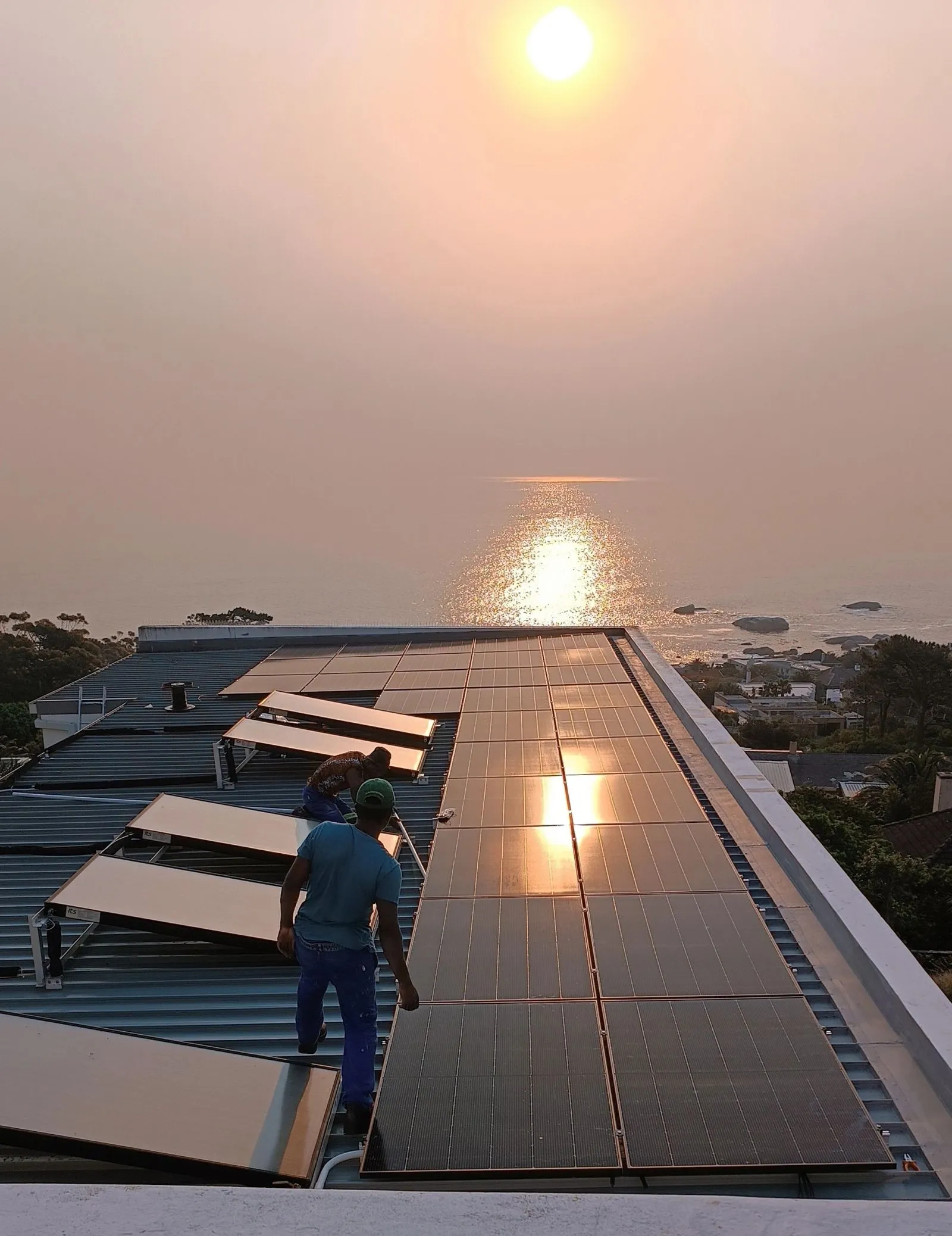 Installers working on a rooftop solar array at sunset, with the sea and coastline below.